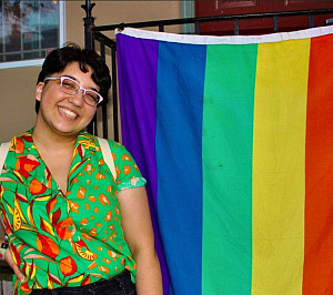 Photograph of a non-binary person standing next to a pride flag.